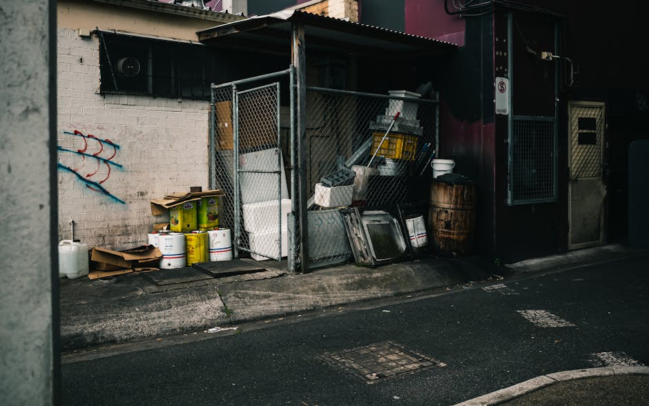 An outdoor area adjacent to a building shows a small enclosed storage space with a metal fence, containing various cleaning equipment and supplies. There are stacked paint cans with yellow and white l